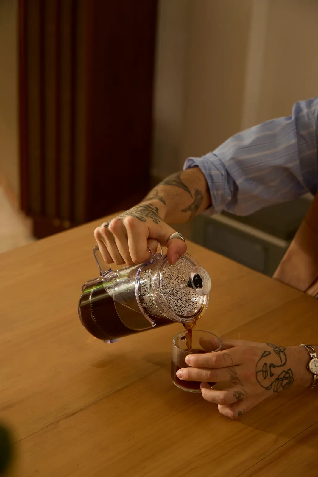 Tattooed hands pouring coffee from a glass pot into a cup on a wooden table