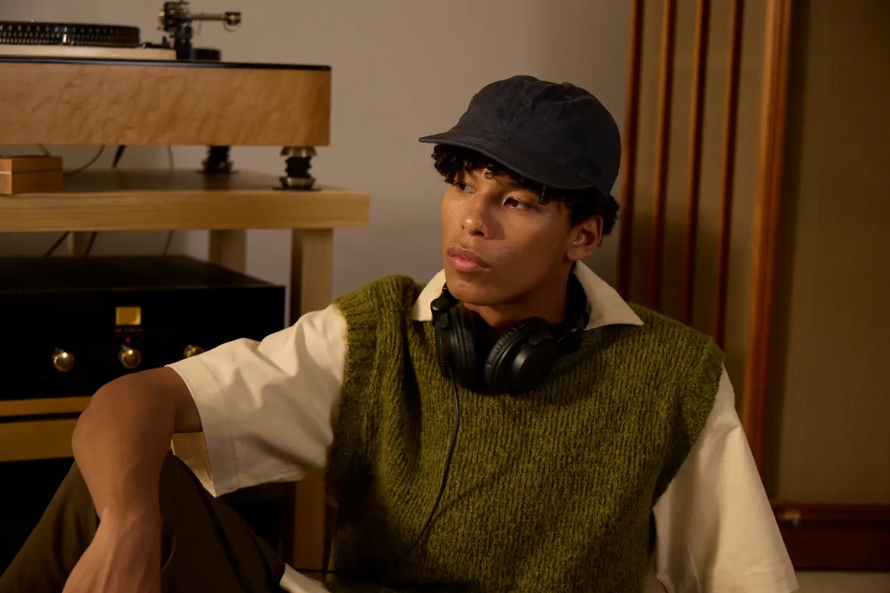 Seated person wearing a cap with headphones around their neck beside audio equipment